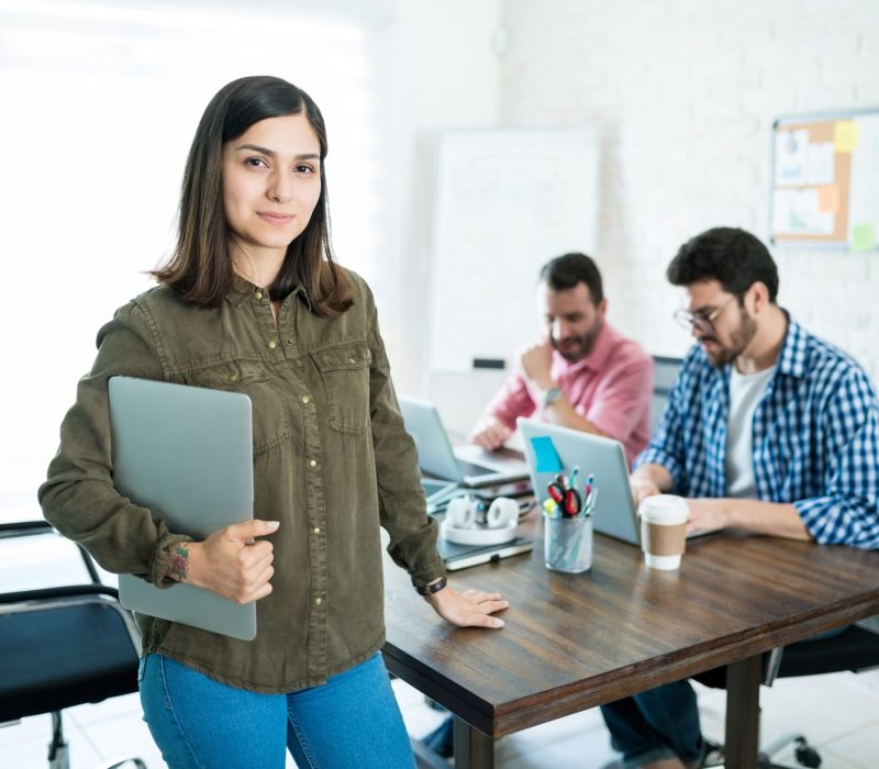 Confident businesswoman with laptop standing against male colleagues in meeting room at office